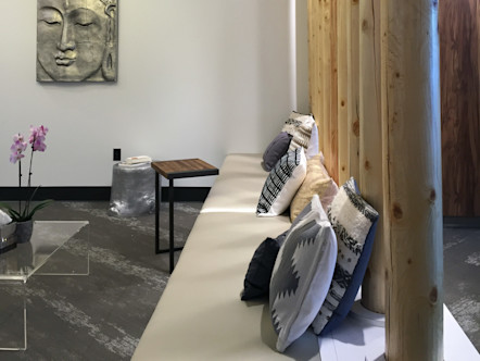 Lobby area with a cream cushioned bench with large light tan wooden poles on its backside with patterned grey toned carpet beneath