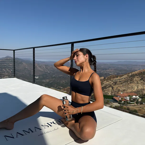Influencer Yaz G in black workout set sitting on white platform with water bottle, overlooking mountainous landscape view