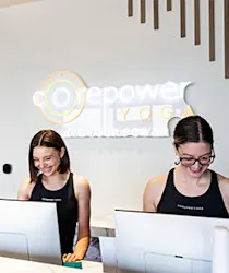 Two women in black tank tops smile while working at computers at a reception desk with a logo on the wall.