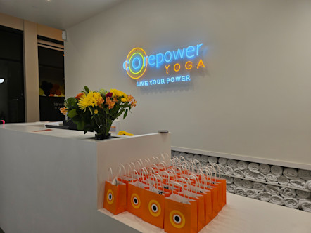 CPY front desk area with a neon CorePower Yoga sign behind a large white desk with flowers on top and a cubby below with rolled up white towels