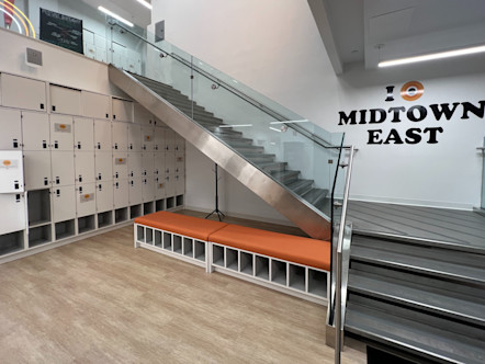 Midtown East CPY logo on white wall behind slate grey stairs with glass railing leading to downstairs area with orange cushioned benches, white lockers, and tan wooden flooring