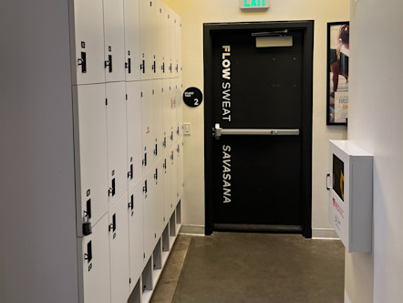 Double row of white lockers on the left hand side of a hallway leading to a black exit door with the words, 'Flow, Sweat, and Savasana' 