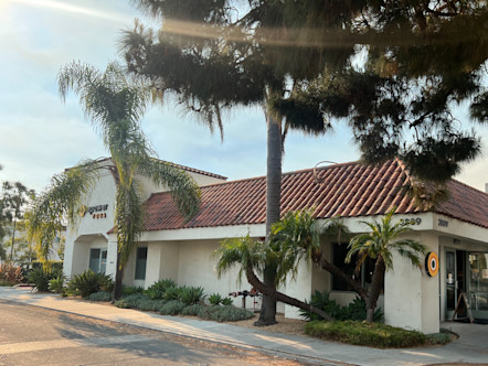 CPY La Cumbre studio with creme-colored stucco and a red brick Spanish style roof, palm trees and greenery surrounding