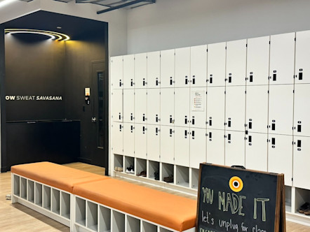 White lockers directly adjacent to a black wall with the words 'flow, sweat, and savasana' to the left and a couple benches with orange cushions in front