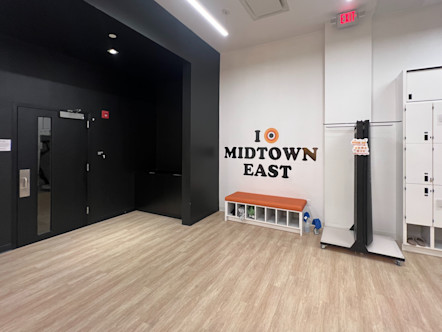 Midtown East CPY logo on a white wall with an orange cushioned bench beneath it directly to the left of a black wall and studio entrance door