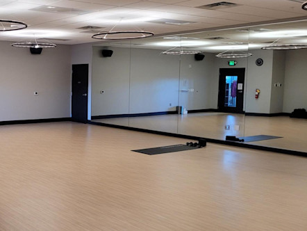 CorePower Yoga studio room with light tan wooden floors, floor to ceiling front mirrors, and a singular black CPY mat for the instructor in the center front of the room