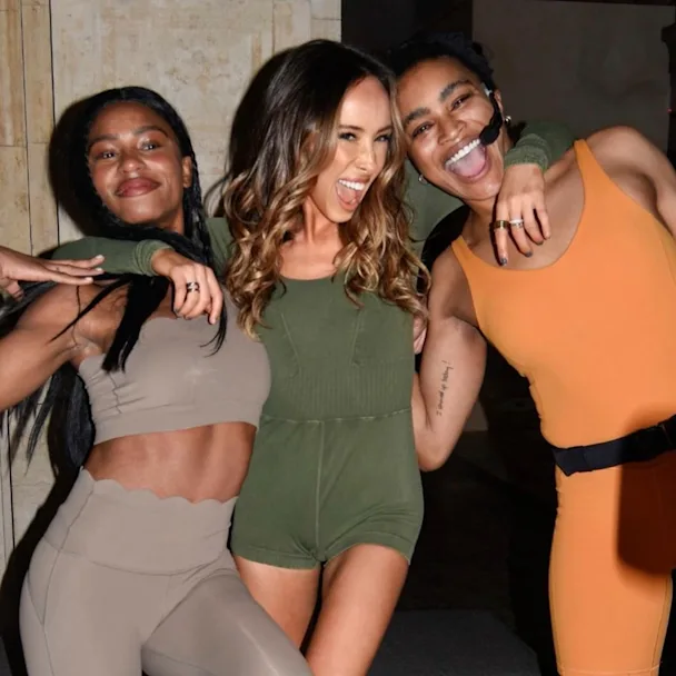 Liv Schreiber in a green onesie smiling and posing with her arms around two CorePower Yoga teachers