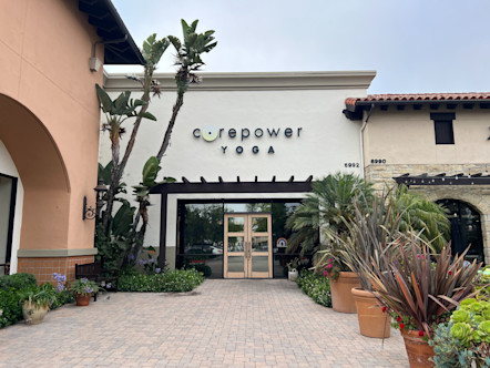 Black CorePower Yoga sign on the front of a large white building with wooden and mirrored double doors and a plant-lined walkway