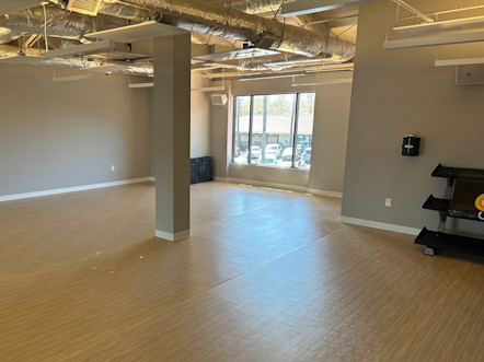 Naturally lit yoga studio with tan beige wooden floors and greige walls with a large window in the back