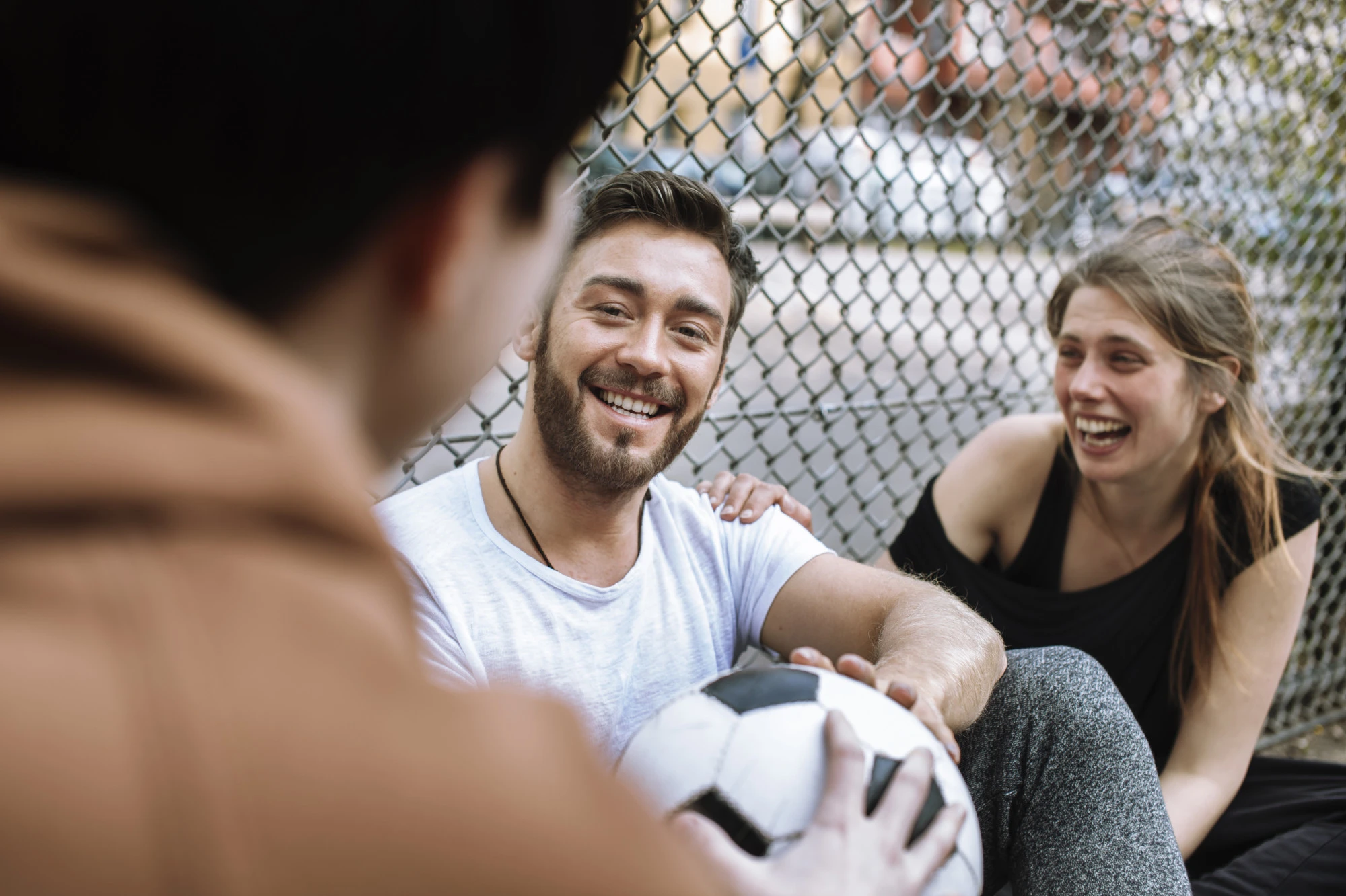 group of 3 people sitting and talking leaning against a fence with a soccer ball