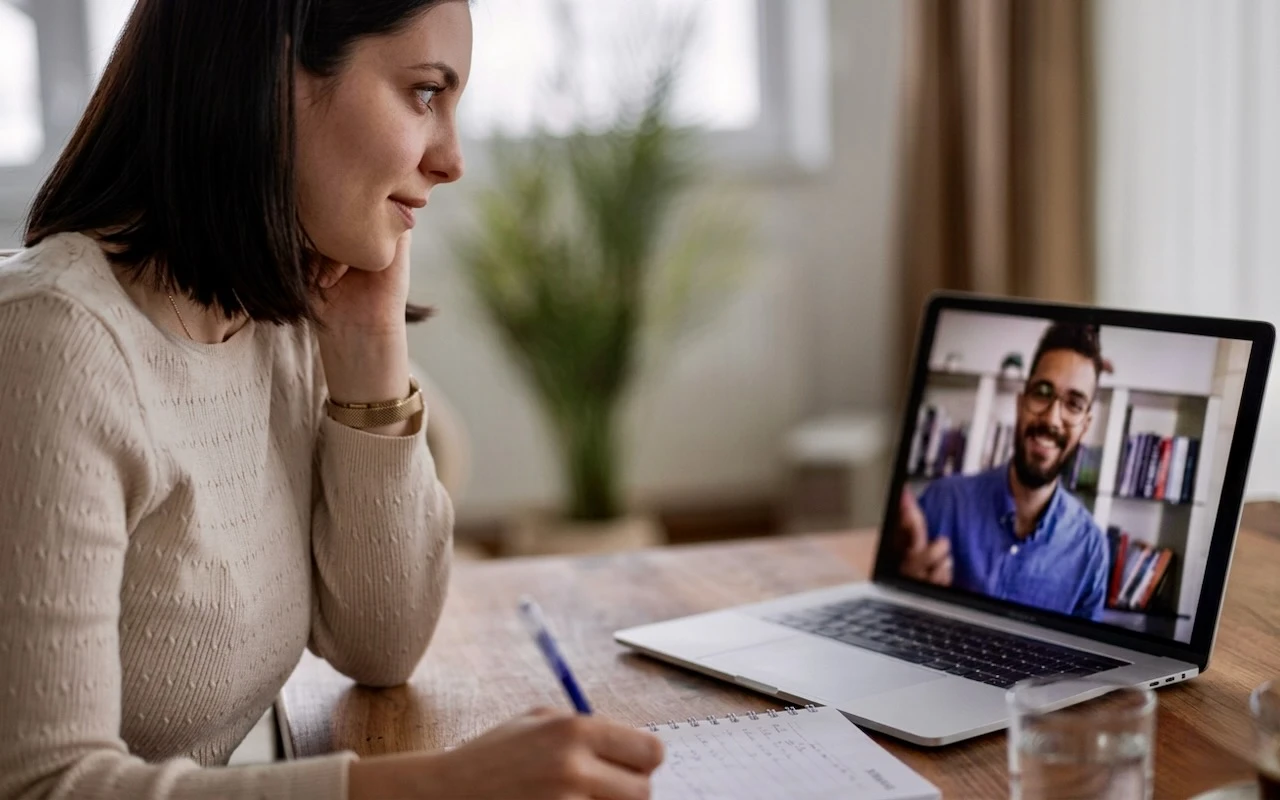 Woman in cream sweater on video call with a man, taking notes at wooden desk with houseplant in background.