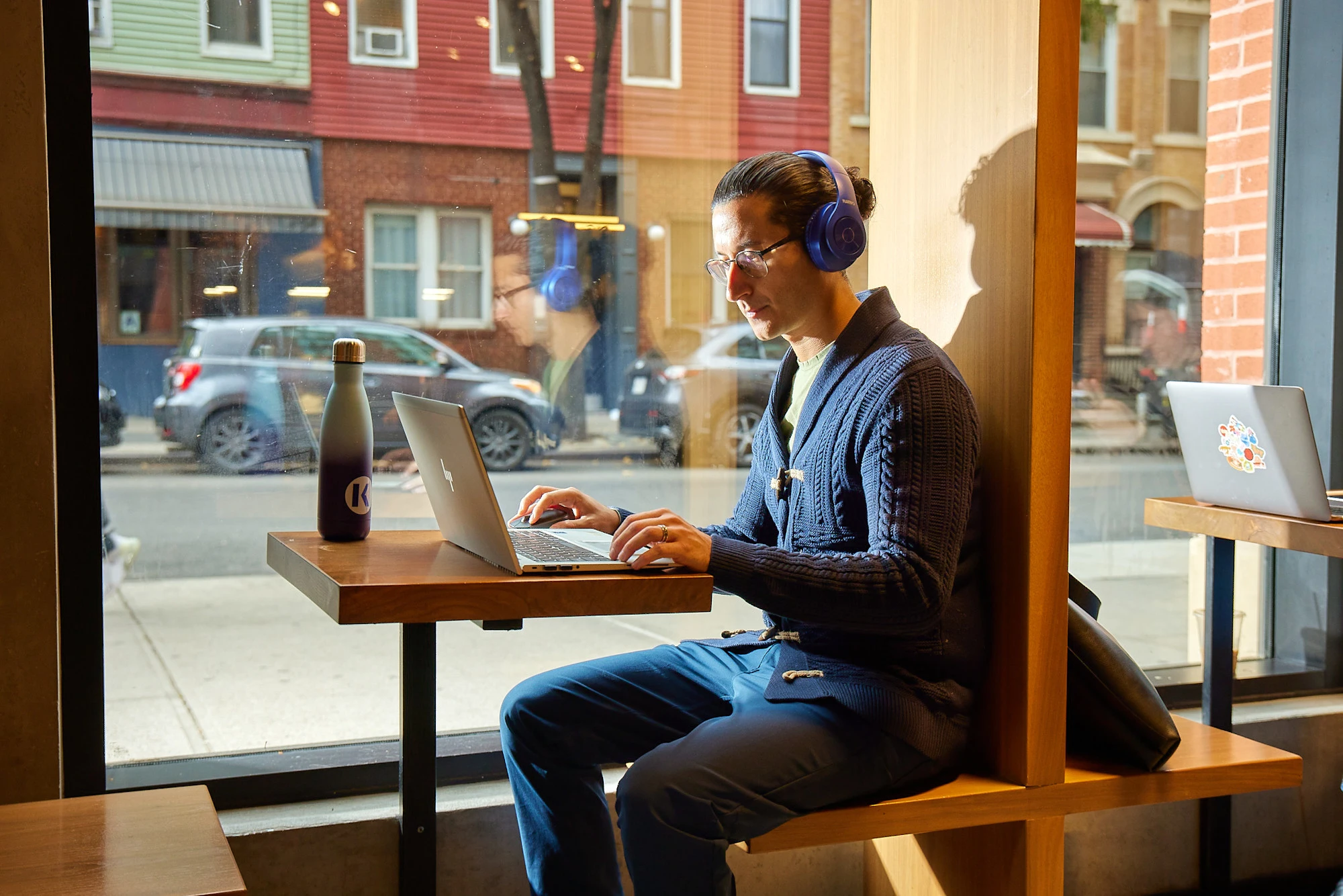 Man with headphones sitting in coffee shop window seat, working on his laptop.