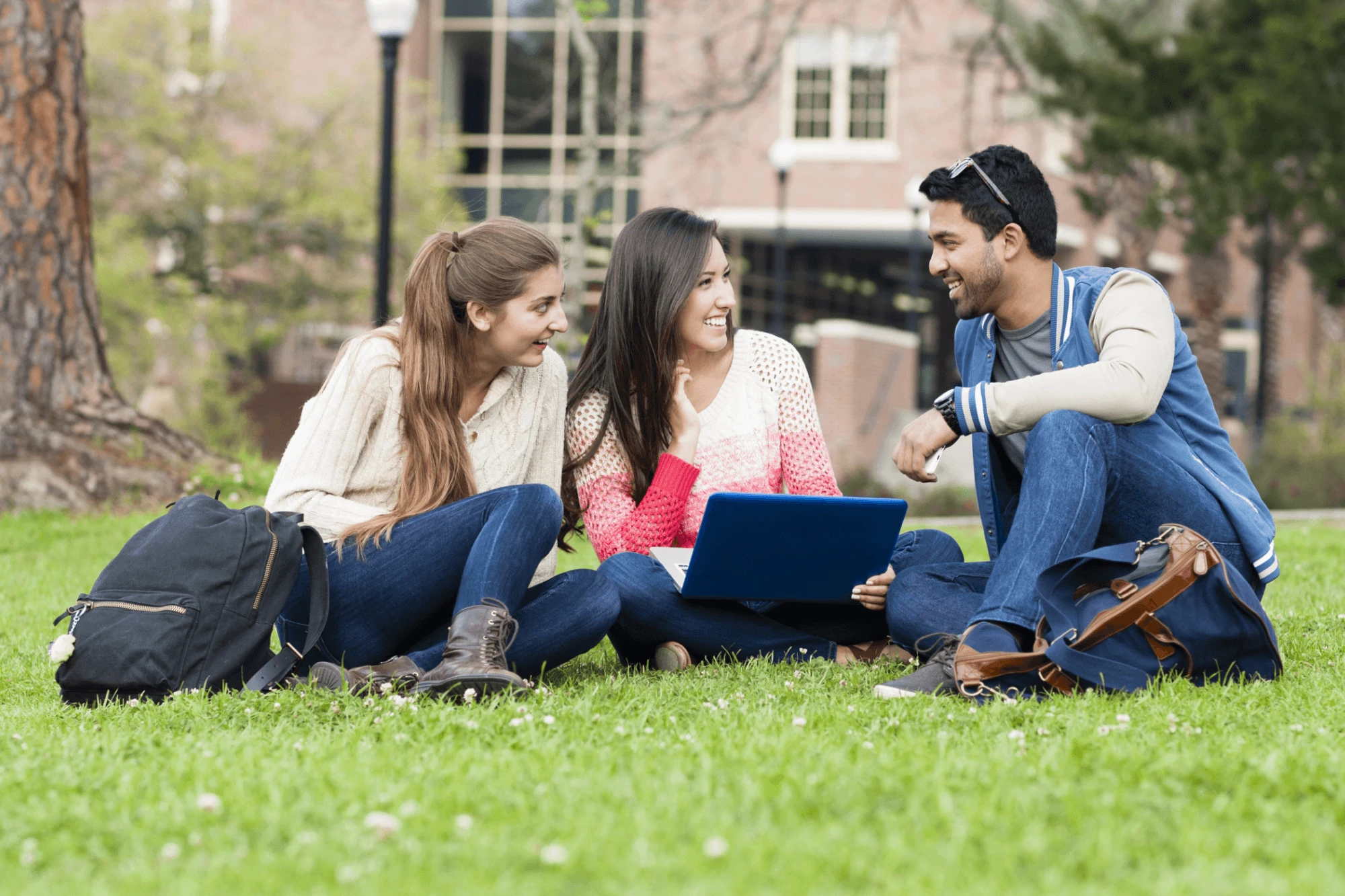 Three students with backpacks sitting on campus lawn, smiling and talking