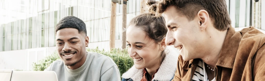 Three smiling students seated outdoors in front of a laptop