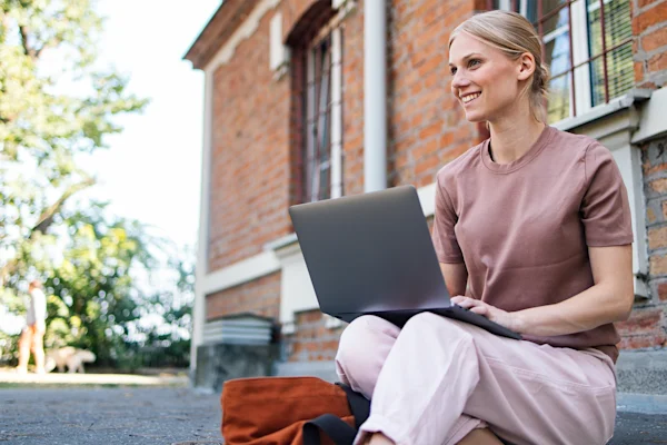 Smiling university student sitting with laptop outside school building
