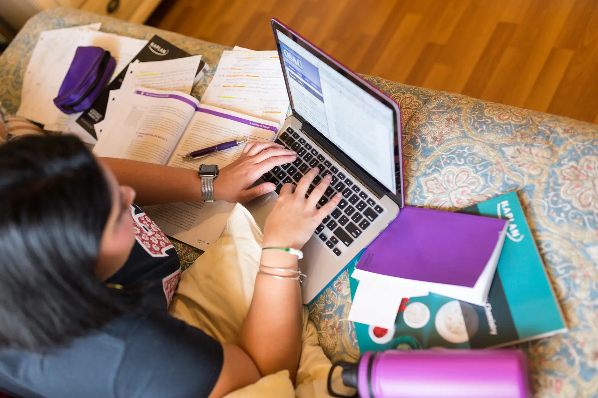 Aerial view of a woman sitting on a carpet over hardwood floors typing on laptop that is on top of a few Kaplan books. 