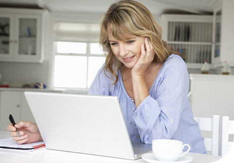 Etwa 50-jährige, blonde Frau mit blauer Bluse, in einer weißen Küche sitzend, die auf ihrem Laptop Informationen zu Prothesenstomatitis sucht.