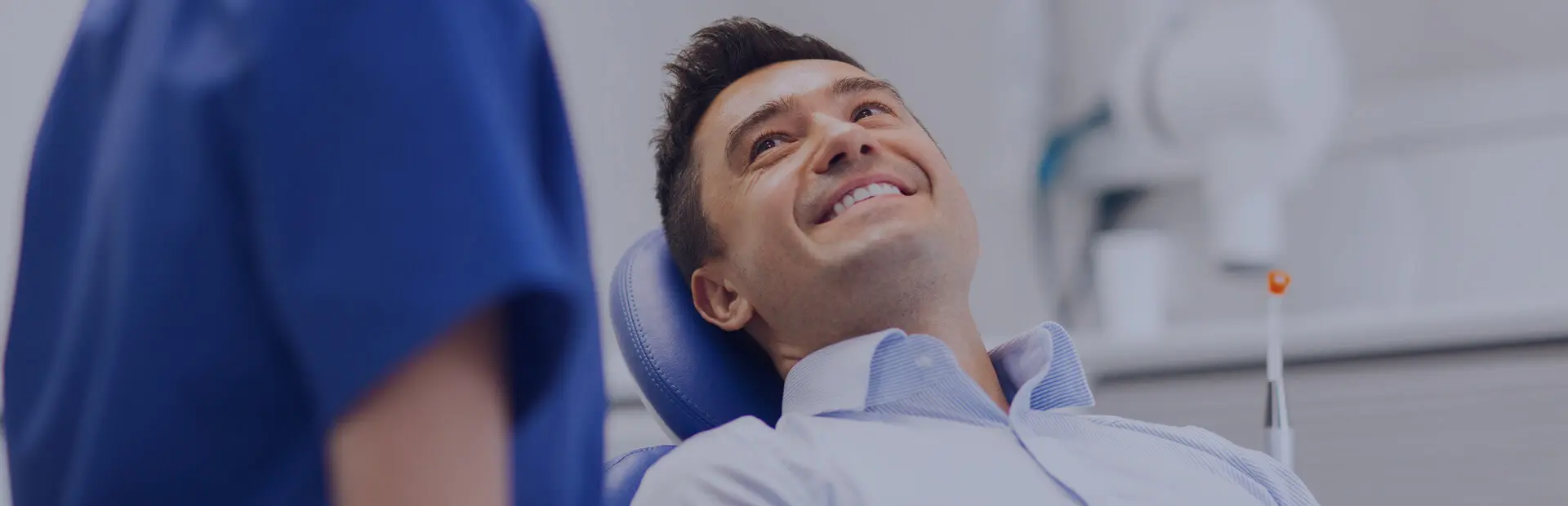 Ein Mann in den 30ern lächelt während einer Kontrolluntersuchung auf dem Zahnarztstuhl, während sein Zahnarzt ihm erklärt, dass blend-a-dent Haftcreme helfen kann, Prothesenwunden zu reduzieren.