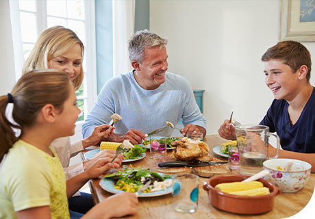 Ein Elternpaar sitzt mit seinen beiden Kindern um einen Tisch. Sie essen Hühnchen und gegrillte Maiskolben. Der Vater lächelt und isst zufrieden, obwohl er herausnehmbaren Zahnersatz hat – dank Fixodent. 
