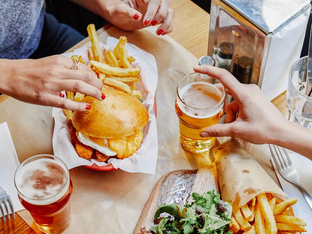 A tabletop setting with two people grabbing for their burger, fries, and beer