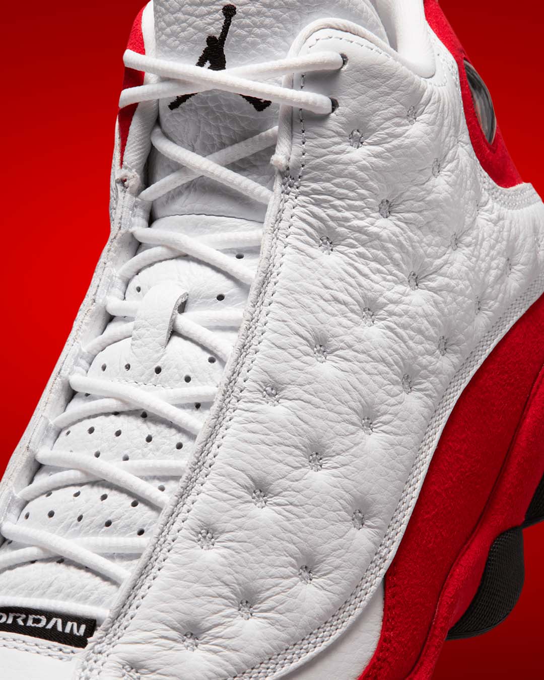 Three-quarter front view of Air Jordan 13 Retro True Red sneaker displaying white quilted tumbled leather upper, red suede toe cap and side panels, black Jumpman logo on tongue, circular hologram detail on ankle, and white midsole with red accents on red studio background
