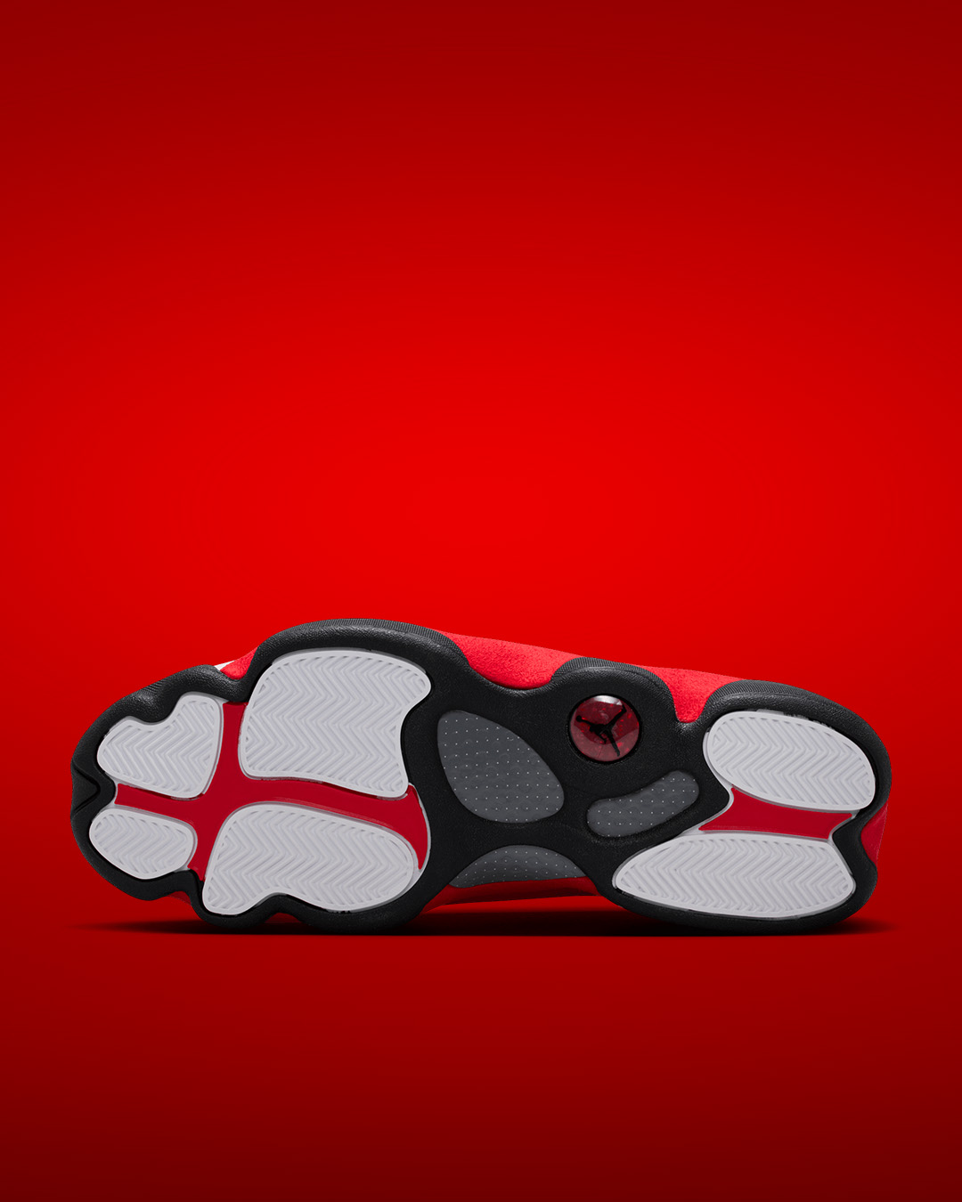 Bottom view of Air Jordan 13 Retro True Red sneaker showcasing paw-inspired black rubber outsole with white herringbone traction pods, grey perforated flex grooves, red Jumpman logo medallion at heel, and red suede upper visible on vibrant red studio background