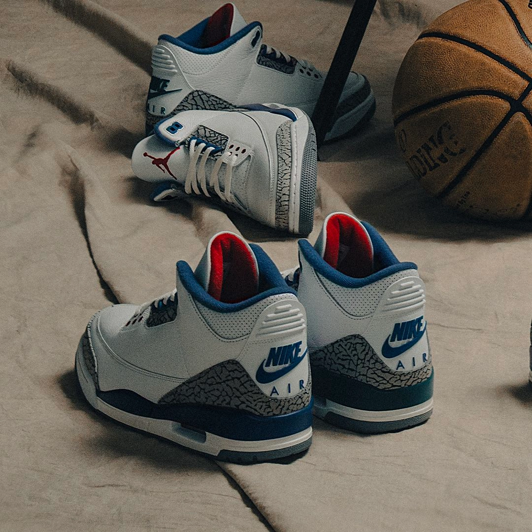 Rear view of two pairs of Air Jordan 3 Retro OG 'True Blue' sneakers showing the Nike Air heel branding, red lining and elephant print midsole, placed on a textured surface next to a basketball