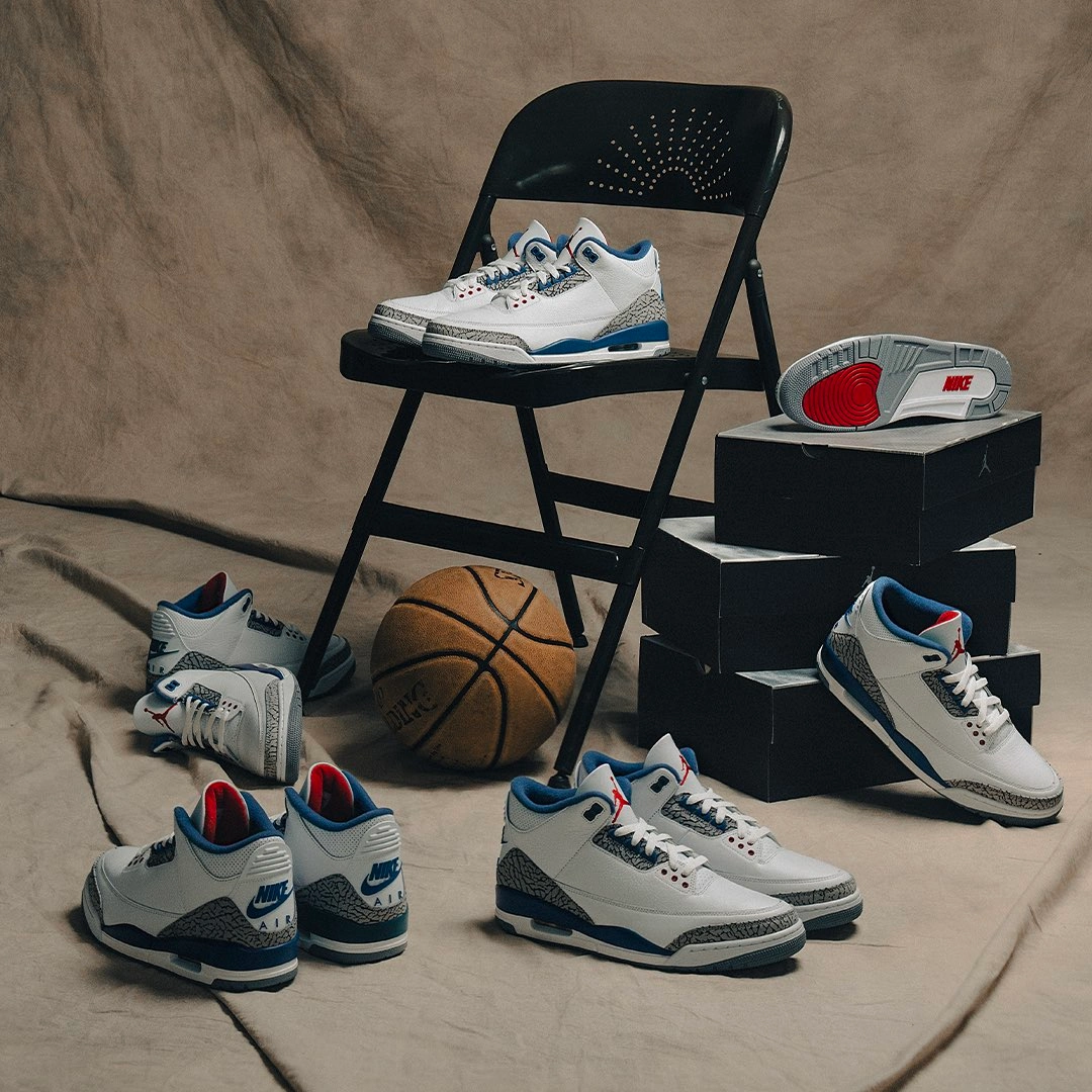 Multiple pairs of Air Jordan 3 Retro OG 'True Blue' sneakers in various sizes arranged on the floor, a black folding chair and stacked shoe boxes, with a basketball in the background, showcasing the full size range