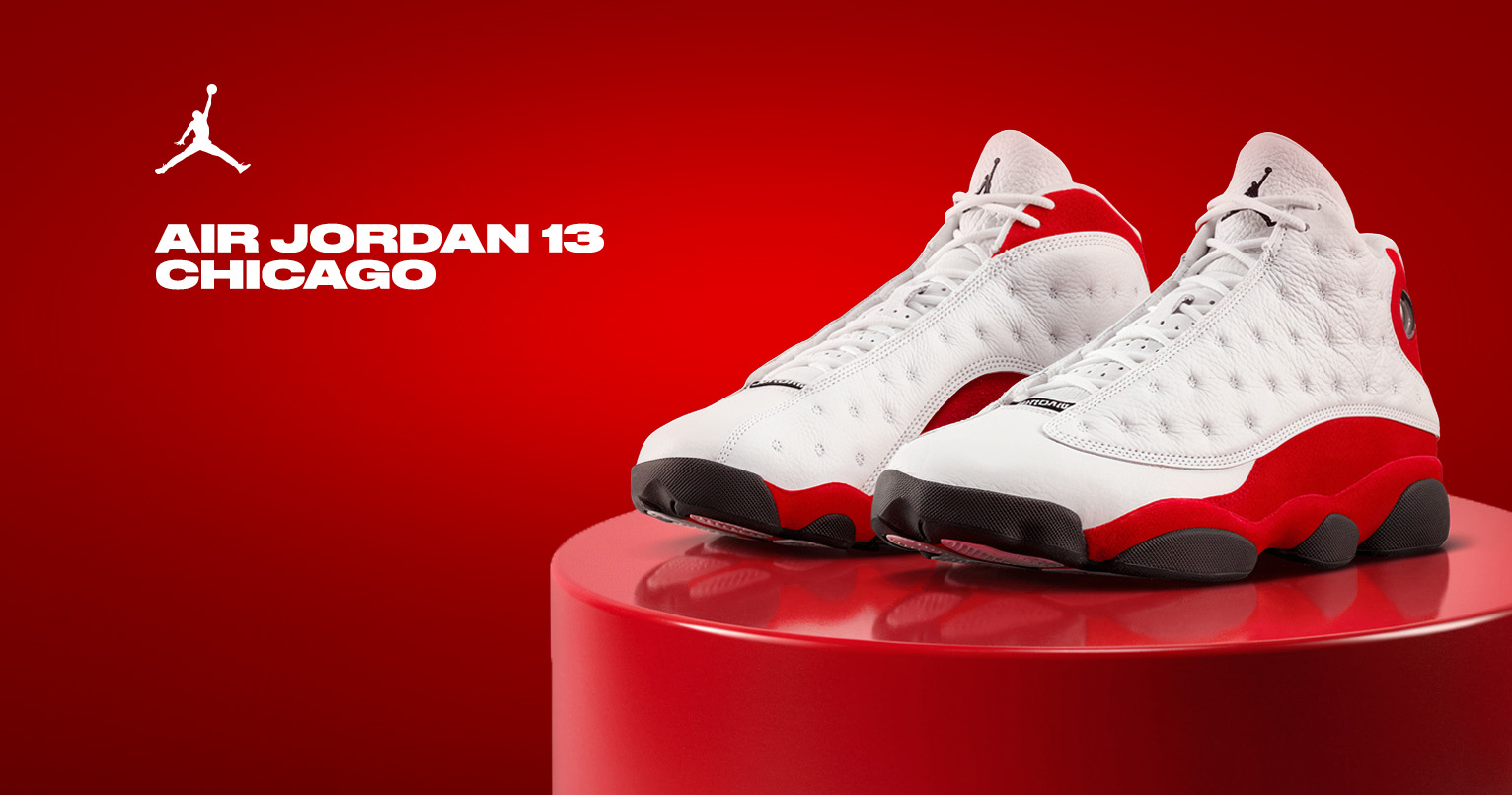 Side profile of Air Jordan 13 True Red sneakers in white quilted leather with bold red outsole and heel counter, black rubber pods, red hologram detail on ankle, and black Jumpman logo on tongue, displayed on glossy red podium against vibrant red background with white Jumpman logo and "AIR JORDAN 13 CHICAGO" text
