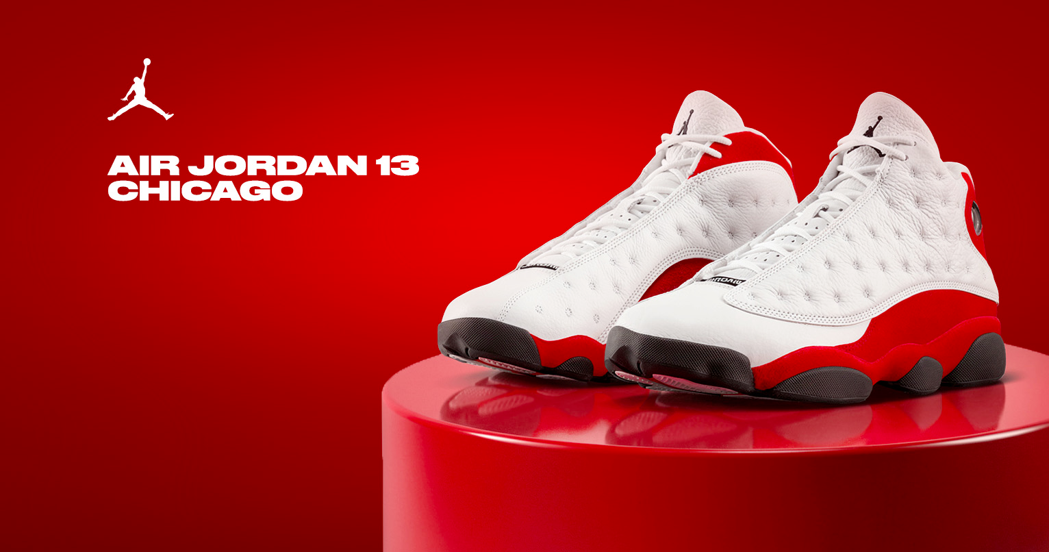 Side profile of Air Jordan 13 True Red sneakers in white quilted leather with bold red outsole and heel counter, black rubber pods, red hologram detail on ankle, and black Jumpman logo on tongue, displayed on glossy red podium against vibrant red background with white Jumpman logo and "AIR JORDAN 13 CHICAGO" text