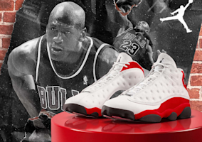 Pair of Air Jordan 13 Retro True Red sneakers with white quilted leather upper, red suede panels and black outsole displayed on glossy red podium against black and white mural featuring Michael Jordan in Chicago Bulls number 23 jersey and white Jumpman logo on brick wall background