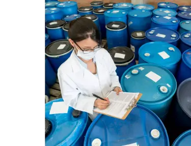 Una mujer escribiendo en un portapapeles rodeada de bidones de productos químicos