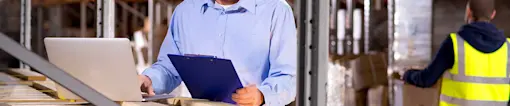 Man with laptop and clipboard standing near pile of empty crates in warehouse