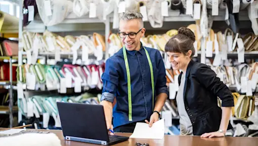 Tailor/designer looking at laptop in warehouse with a lot of fabric rolls in the background.