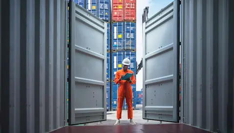 Man in work clothes taking notes while looking into empty container in container shipyard.