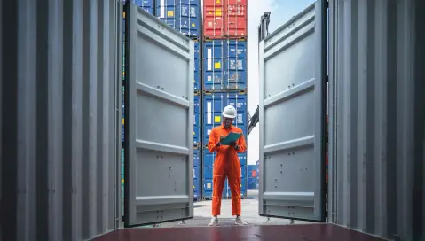 Man in work clothes taking notes while looking into empty container in container shipyard.