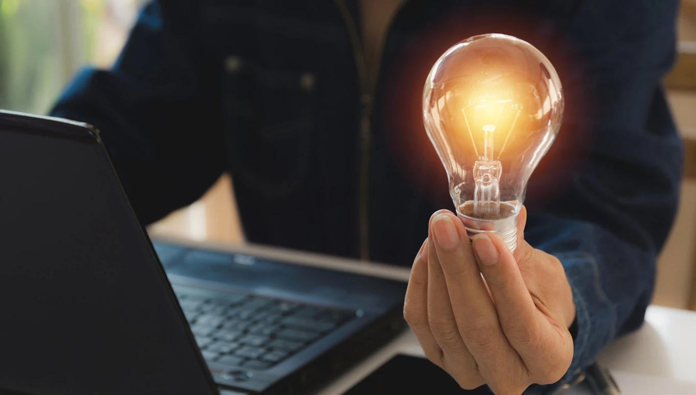 Person sitting in front of computer and holding a light bulb in hand that glows.