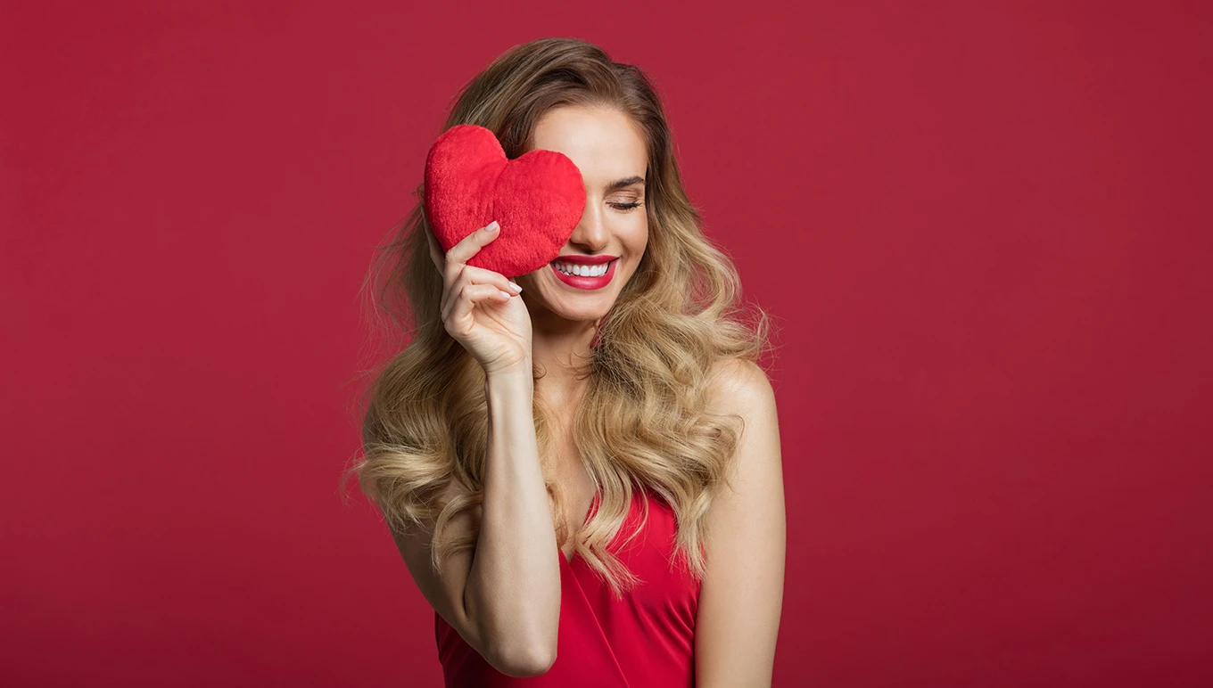 Woman in red dress smiling as she holds stuffed heart to face