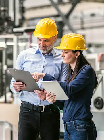 Two factory workers in hard hats reviewing tablet and clipboard.