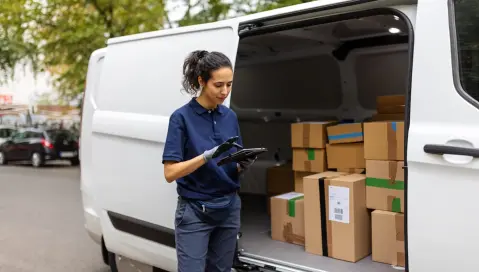 Woman standing by van with parcels and looking at tablet.