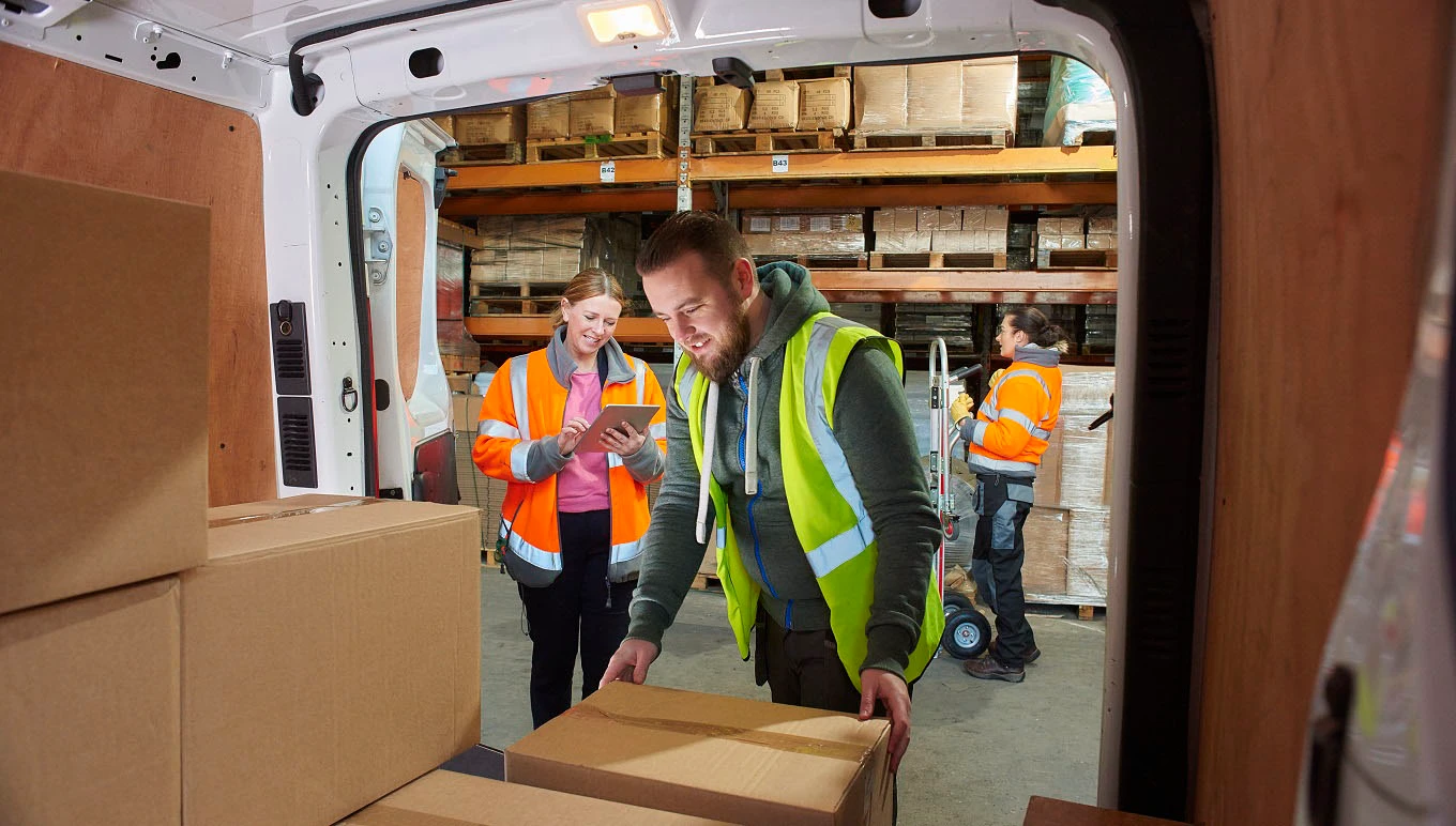 Warehouse worker loading delivery van while another takes inventory on tablet.