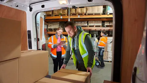 Warehouse worker loading delivery van while another takes inventory on tablet.