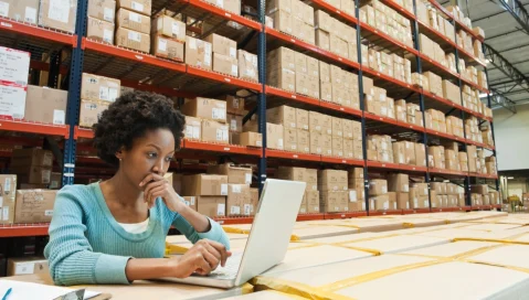 A manufacturing and distribution professional considers data on a laptop with a warehouse of supplies in the background.