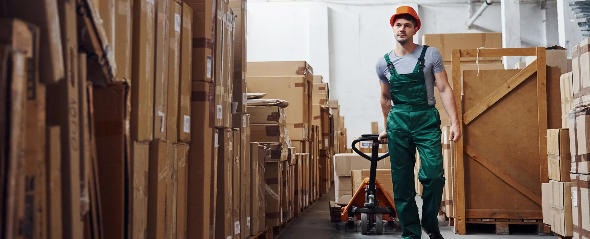 Warehouse worker moving pallet jack through aisle of boxed inventory