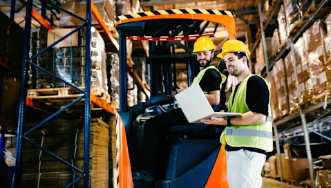 Two workers in hardhats using a laptop near forklift