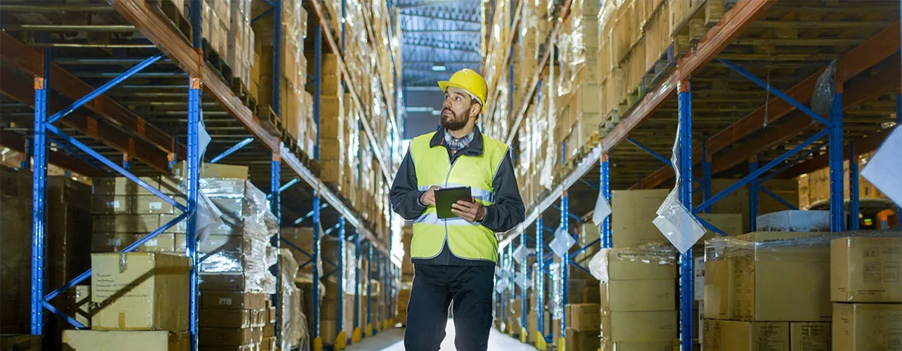 A man wearing a hard hat walking around a warehouse and looking at the shelves