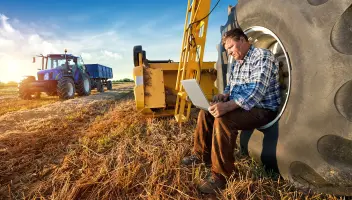 Farmer sitting on large tractor wheel looking at laptop