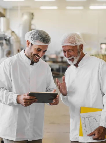 Two men in protective outfits looking at tablet on food factory shopfloor.