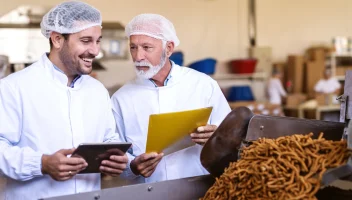 Two food factory workers review a document and use a tablet.