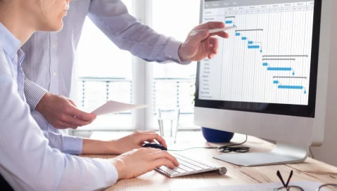 Person typing on a desktop next to a colleague pointing at charts on screen